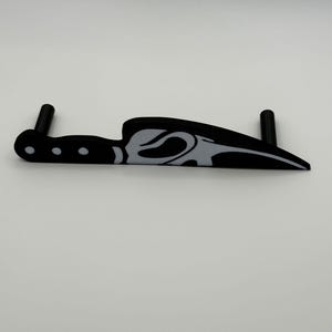 May include: A black and white decorative knife with a curved blade and a handle with three small circles. The knife has a stylized design on the blade and two black cylindrical posts extending from the handle.