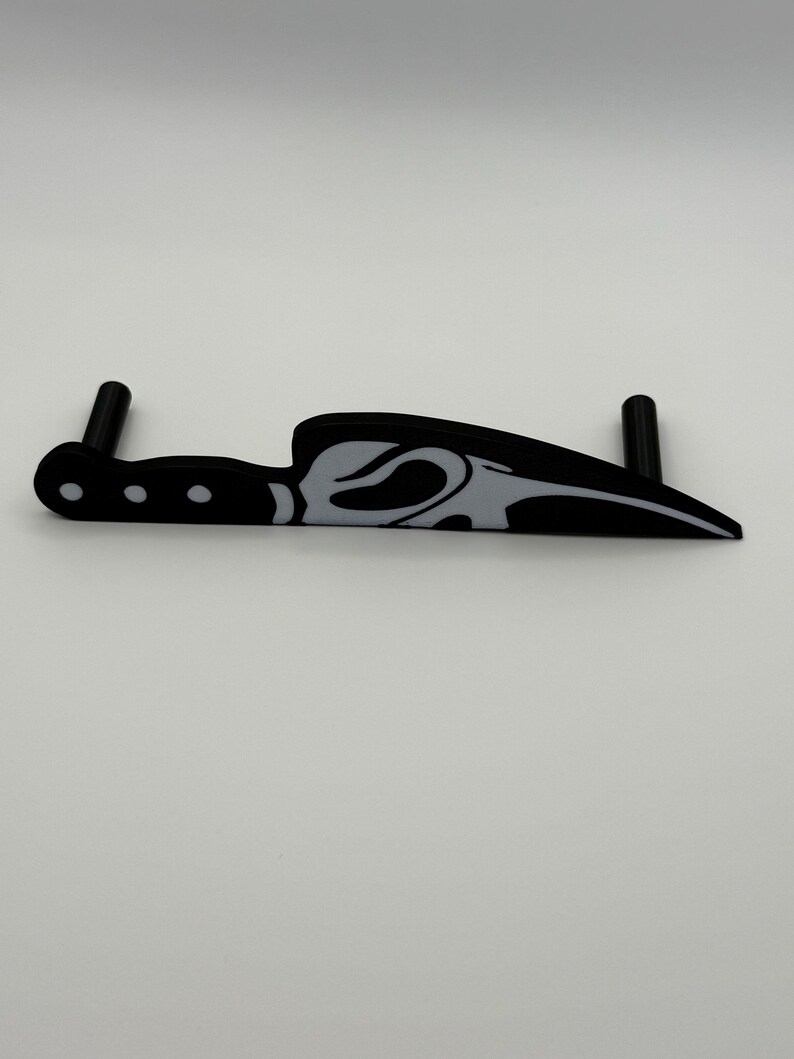 May include: A black and white decorative knife with a curved blade and a handle with three small circles. The knife has a stylized design on the blade and two black cylindrical posts extending from the handle.