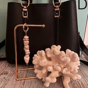 May include: A black handbag with gold-tone hardware, a gold-tone metal display stand, and a keychain featuring stacked white and grey marbled stone beads with a heart-shaped pendant. A white coral-like decorative object sits in the foreground.