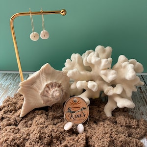 May include: Gold hoop earrings with small, round, off-white shell charms. One pair hangs from a gold metal stand, while another pair is attached to a circular cardboard tag labeled Kini's Collection with a shell graphic and sun icon.