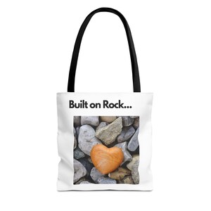 Könnte beinhalten: Weiße Tragetasche mit schwarzen Griffen. Auf der Tasche steht der Text "Built on Rock...". Das Design zeigt eine Nahaufnahme verschiedener Steine, mit einem herzförmigen Stein in der Mitte.