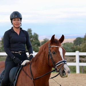 May include: A person in a black riding helmet and attire sits on a chestnut horse. The horse has a white stripe on its face and a jeweled browband. The background features a white fence and trees under a cloudy sky.