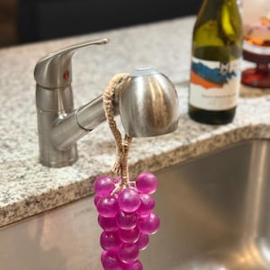 May include: A purple grape-shaped soap hanging from a kitchen tap. The soap is made of small, round, translucent purple beads. The tap is stainless steel, and a bottle of wine is in the background.