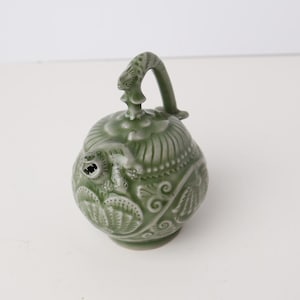 Könnte beinhalten: Ein grüner Keramik-Räuchergefäß mit Griff und aufwendigen Blumen- und Blattmustern. Der Brenner hat einen runden Körper mit einer kleinen Öffnung und steht auf einer weißen Oberfläche.