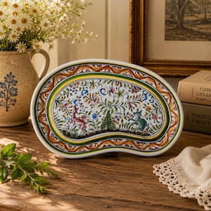 May include: A kidney-shaped ceramic platter featuring a hand-painted design of colorful flowers, green foliage, and two small animals. The border consists of concentric rings in green, yellow, and brown with a wavy, rope-like pattern on the outer edge.