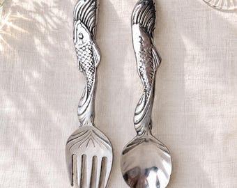 May include: A silver-toned metal fork and spoon set featuring handles shaped like fish with detailed scales and fins. The fork has four tines, and the spoon has a smooth, rounded bowl. Both utensils have a polished, reflective finish.