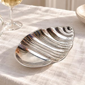 May include: A silver-toned metal dish shaped like a seashell with a smooth, polished finish and deep, curved ridges along one side. The dish has an elongated, oval form with a slightly pointed end, reflecting light across its metallic surface.