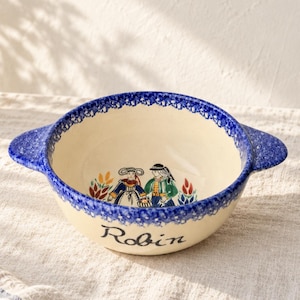 May include: A cream-colored ceramic bowl with two side handles, featuring a blue speckled rim and a hand-painted illustration of two figures in traditional attire. The name Robin is written in black script on the front of the bowl.