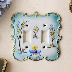 May include: A white ceramic double light switch plate with a scalloped edge, featuring a hand-painted scene of trees with yellow blossoms and a person sitting on a blue bench under a yellow umbrella, accented with light blue borders and gold trim.