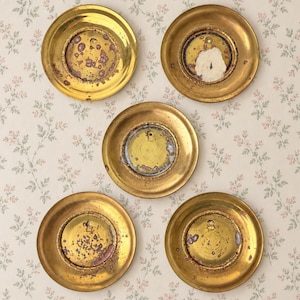 May include: Five circular brass wall plates arranged in a pentagon pattern. Each plate features a wide, polished gold-toned rim and a recessed center with visible oxidation, dark spots, and remnants of adhesive or paper backing in the middle.