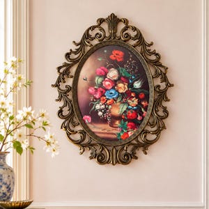 May include: An oval wall art piece featuring a colorful floral still life painting in a gold-toned, ornate, scrolled metal frame. The painting depicts a bouquet of red, blue, yellow, and pink flowers in a vase against a dark background.