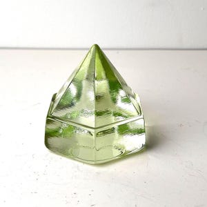 Op de afbeelding: Een kleine, geometrische, lichtgroene glazen doos met een piramidevormig deksel. De doos heeft een facetontwerp en het glas lijkt enigszins doorschijnend. De doos staat op een wit oppervlak.