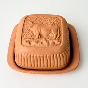 May include: A terracotta butter dish with a cow relief design on the lid. The dish is rectangular with a ridged base and a matching tray. The cow is depicted in a field, adding a rustic touch to the kitchenware.