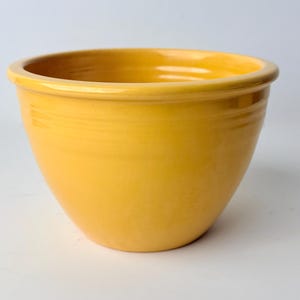 Puede incluir: Un cuenco mezclador de cerámica de color amarillo brillante con un acabado liso y brillante. El cuenco presenta un borde ancho y redondeado y una serie de tres anillos concéntricos horizontales en relieve alrededor de la parte superior.