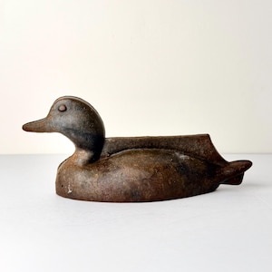 May include: A weathered, brown cast iron duck figurine. The duck is in a resting position, with its head upright and beak forward. The figurine has a rustic, aged appearance, with visible texture and color variations.