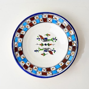 May include: A round, white ceramic plate with a blue rim and a decorative border of brown, blue, and gold squares. The center features a symmetrical design of two birds and floral elements in green, red, and blue.