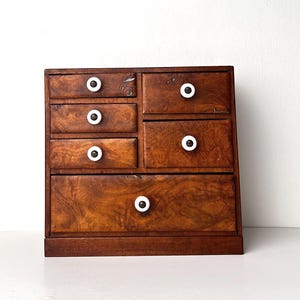 May include: A small, wooden chest of drawers with six drawers of varying sizes. The wood has a rich, warm brown tone with visible grain. Each drawer is accented with a white, circular knob. The chest sits on a flat base.