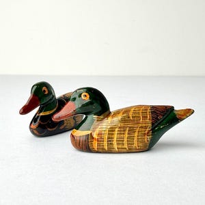 May include: Two hand-painted wooden duck figurines. The ducks have green heads, red beaks, and brown and yellow bodies. One duck is in front of the other. The background is white.