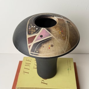 Puede incluir: Un jarrón de cerámica negro y beige con diseños geométricos, incluyendo formas rosas y marrones. El jarrón está sobre dos libros, uno amarillo y otro rojo, con las palabras "TIME FOR POETRY" visibles.