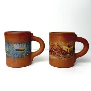May include: Two terracotta mugs with brown handles. One mug features a blue and yellow design, while the other has a brown and yellow pattern. The mugs are displayed against a white background.