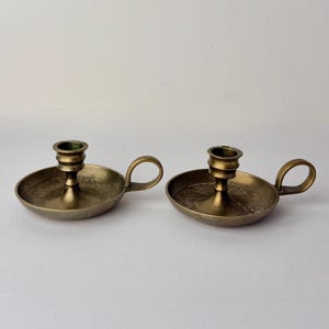 May include: Two antique brass candle holders. Each holder has a circular base with a small handle and a central cup to hold a candle. The brass has a warm, aged patina, suggesting vintage or antique origins. The design is simple and functional.