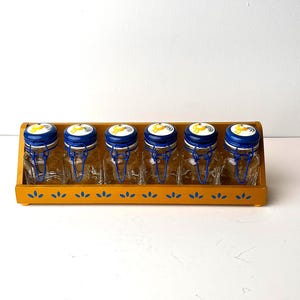 Puede incluir: Un conjunto de seis pequeños tarros de cristal con tapas azules y blancas, asegurados con cierres de alambre. Los tarros se exhiben en un estante de madera amarilla con un patrón floral decorativo. Las tapas tienen un diseño amarillo. Los tarros están vacíos.