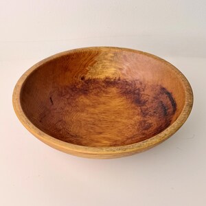 May include: A wooden bowl with a natural wood grain finish. The bowl is round with a wide, shallow design. The wood has a warm, reddish-brown hue with darker areas, showcasing the natural patterns of the wood.