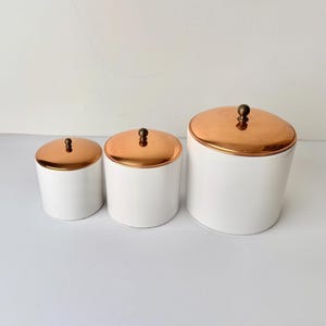 May include: Three white ceramic storage jars with copper-coloured lids and small, round knobs. The jars are arranged in a row, varying in size. The lids have a shiny, metallic finish. Ideal for kitchen storage.