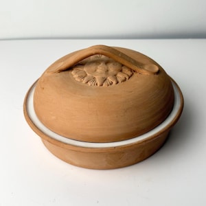 Puede incluir: Un plato redondo de cerámica de color terracota con una tapa a juego. La tapa presenta un asa y un diseño floral. El plato se asienta sobre una base blanca, creando un contraste con los tonos terrosos de la cerámica.