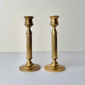 May include: Two gold-coloured candlesticks with a classic design. Each candlestick has a round base, a fluted column, and a cup-shaped top. The candlesticks are made of metal and have a matte finish. They are placed on a white surface.