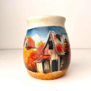 以下が含まれることがあります： 手描きの小さなセラミックジャー。風景画が描かれています。 赤い屋根の納屋が、オレンジ、黄色、茶色の秋の葉に囲まれています。 ジャーはベージュの縁と底があり、背景は青い空です。