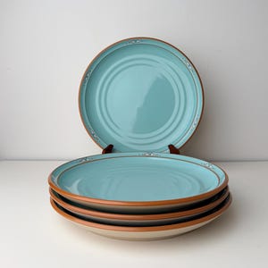 Puede incluir: Una pila de cuatro platos de cerámica turquesa con borde marrón y borde decorativo. Un plato se exhibe en un soporte de madera. Los platos tienen un diseño de círculos concéntricos en el centro.