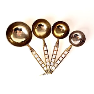 May include: A set of four brass measuring spoons. Each spoon has a round bowl and a handle inlaid with rectangular mother-of-pearl tiles. The spoons are arranged in a fan shape against a white background. The spoons are for measuring dry and liquid ingredients.