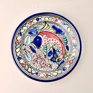 May include: A round, hand-painted ceramic plate with a vibrant fish design. The fish is primarily red and blue, with a dotted pattern. The plate has a dark blue rim and a decorative border with colorful accents.