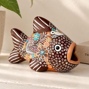 May include: A brown ceramic fish figurine featuring an open mouth and intricate hand-painted details. The body is decorated with white dots, colorful floral patterns, and white line accents on the fins. The eye is white with a black pupil.