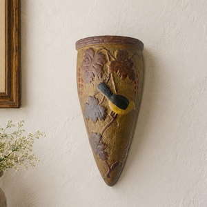 Puede incluir: Una maceta de pared en forma de cono con una superficie texturizada de color verde oliva, decorada con relieves de hojas de vid marrones, bayas rojas y un pequeño pájaro con pecho amarillo y alas negras, con un borde geométrico.