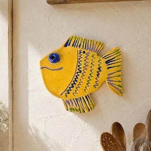 Puede incluir: Una decoración de pared de cerámica amarilla con forma de pez, que presenta un ojo azul, una boca pintada de azul y patrones en zigzag de color azul oscuro y verde en el cuerpo y las aletas. Tiene un pequeño orificio cerca de la aleta superior.