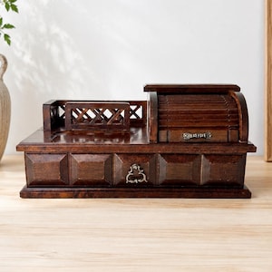 May include: A dark brown wooden desk organizer featuring a roll-top compartment with a small metal handle, a lattice-style side fence, and a front drawer with a decorative metal pull. The piece has a paneled front design and a smooth finish.