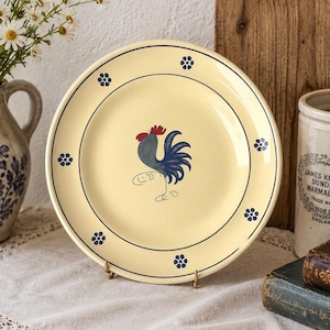 May include: A round, cream-colored ceramic plate featuring a central illustration of a blue rooster with a red comb. The rim is decorated with a thin dark blue circular line and six small, evenly spaced blue flower motifs around the edge.