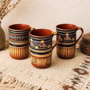 May include: Three cylindrical terracotta mugs featuring hand-painted geometric and floral patterns in black, blue, yellow, and white. Each mug has a sturdy handle and a smooth, reddish-brown interior finish, showcasing detailed craftsmanship.