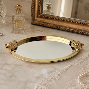 May include: An oval-shaped vanity tray with a polished gold-tone metal frame and a mirrored base. The tray features decorative gold-tone metal floral accents on both sides, serving as handles. The surface is reflective and smooth.
