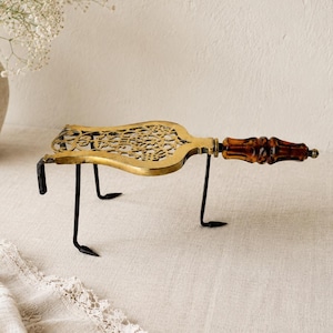May include: A brass trivet featuring an intricate openwork floral pattern, supported by three slender black metal legs. The handle is crafted from a polished, translucent amber-colored material with a segmented, decorative shape.