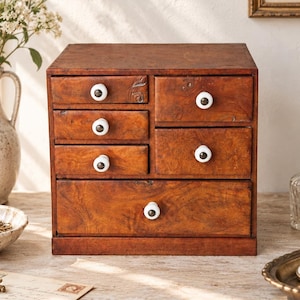 May include: A wooden desktop organizer featuring a rich brown grain finish and six drawers of varying sizes. Each drawer is equipped with a small, round, white ceramic knob. The unit has a rectangular shape with a flat top and a solid base.