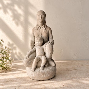 May include: A light gray stone-colored sculpture depicting a seated adult figure with long hair wearing a collared shirt, with a small child sitting on their lap. The figures are rendered in a smooth, matte finish with subtle facial features.