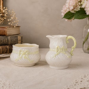 May include: A white ceramic creamer pitcher and a matching sugar bowl, both featuring a subtle swirl texture and a pale yellow ribbon detail molded into the surface. The pitcher has a curved handle and a scalloped rim, while the bowl is round.