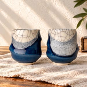 May include: Two ceramic cups featuring a distinct two-tone design with a glossy deep blue base and a cream-coloured upper section. The cream portion displays a prominent dark crackle glaze pattern, while the overall shape is organic and slightly irregular.