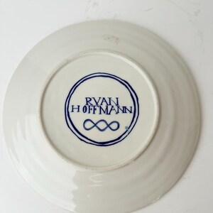 May include: White ceramic plate with a blue circular design on the bottom. The design includes the text "RYAN HOFFMANN" and an infinity symbol. The plate has a slightly ridged edge.