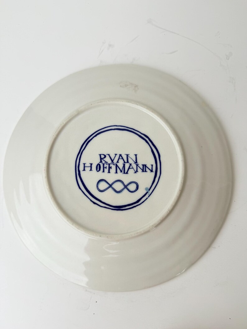 May include: White ceramic plate with a blue circular design on the bottom. The design includes the text "RYAN HOFFMANN" and an infinity symbol. The plate has a slightly ridged edge.