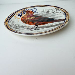 May include: Oval ceramic plate with a detailed illustration of a bird in shades of red, orange, and brown. The plate has a gold and blue border, and the bird is set against a white background with floral accents.