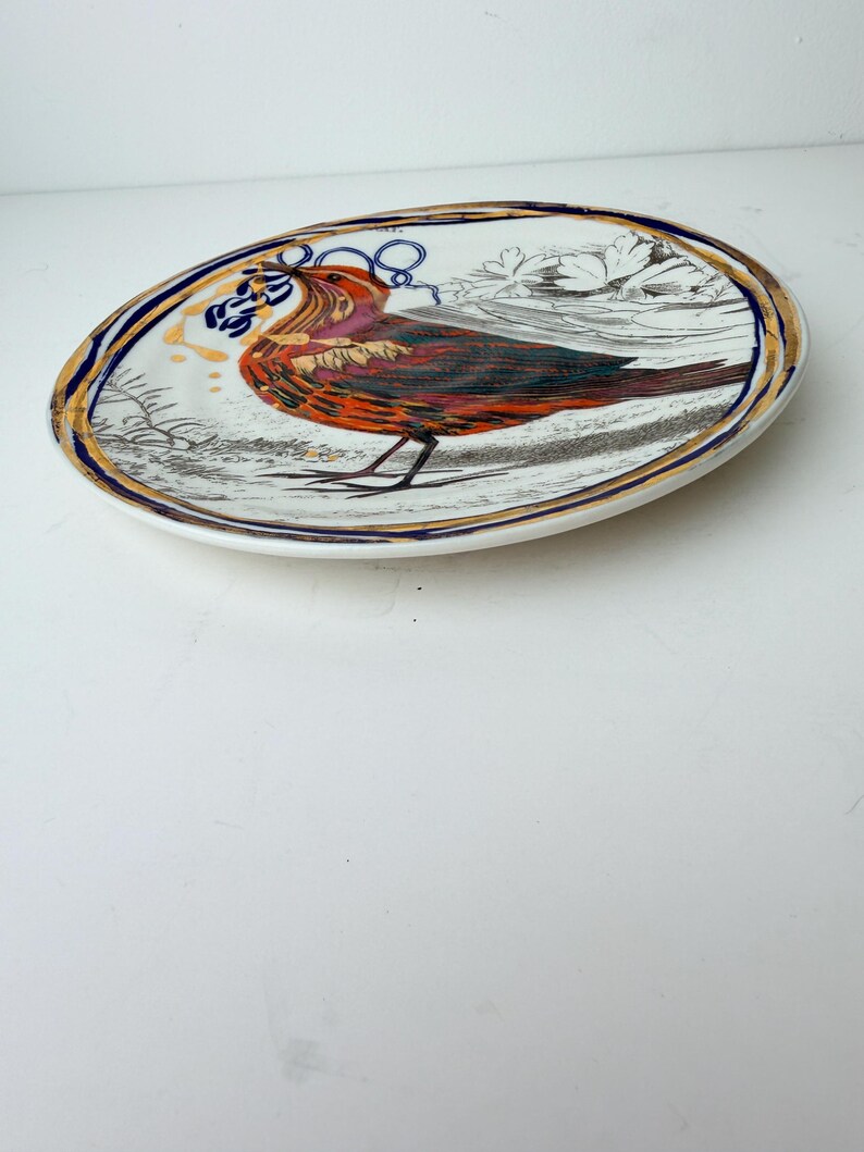 May include: Oval ceramic plate with a detailed illustration of a bird in shades of red, orange, and brown. The plate has a gold and blue border, and the bird is set against a white background with floral accents.